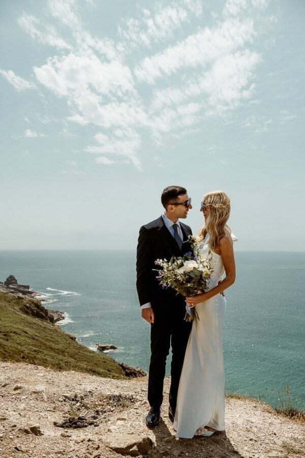 Bride and groom standing together on Cornish clifftop with wildflower bouquet, overlooking the ocean