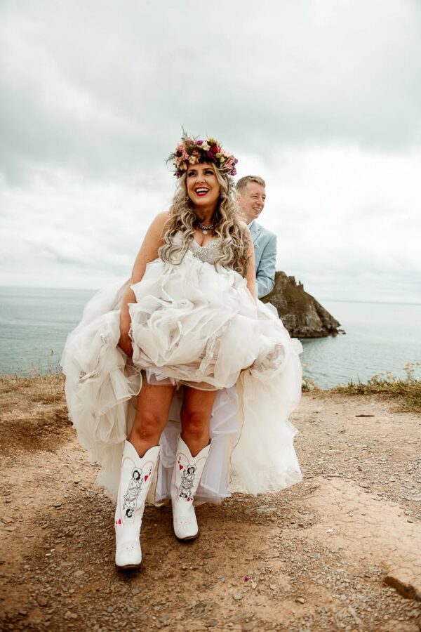 Joyful bride in flower crown lifting skirt to show custom cowboy boots at Torbay Devon wedding
