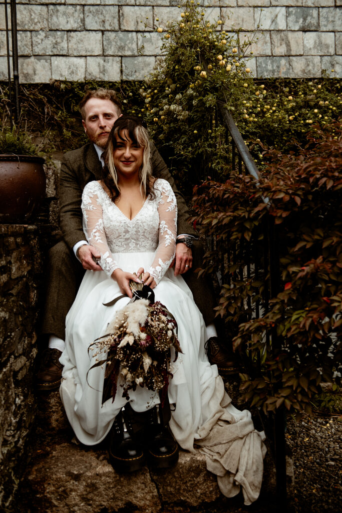 Bride in lace dress sitting against stone wall with dried flower bouquet Wonwood Barton