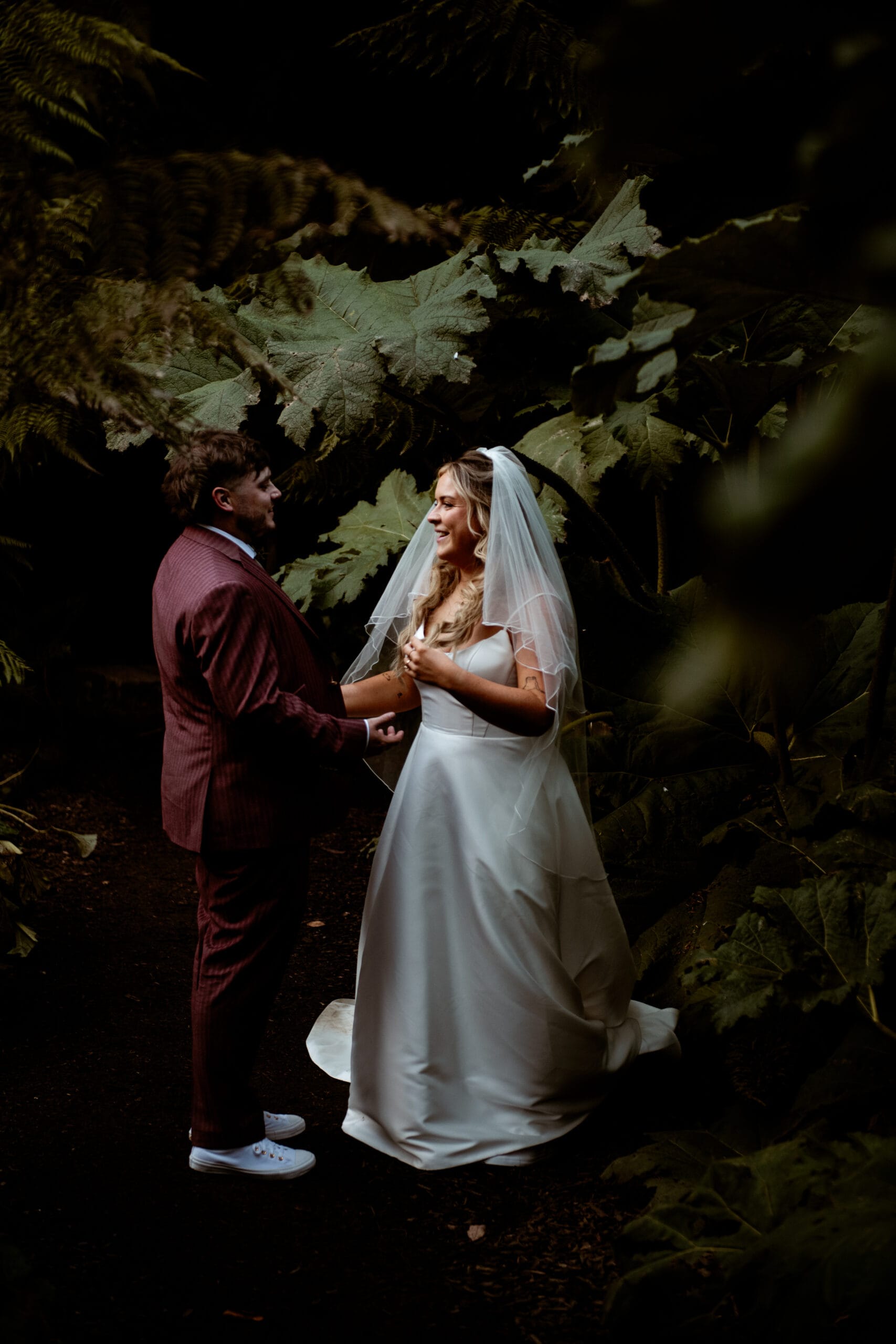 Groom in burgundy suit and bride in veil sharing intimate moment at Heligan garden wedding