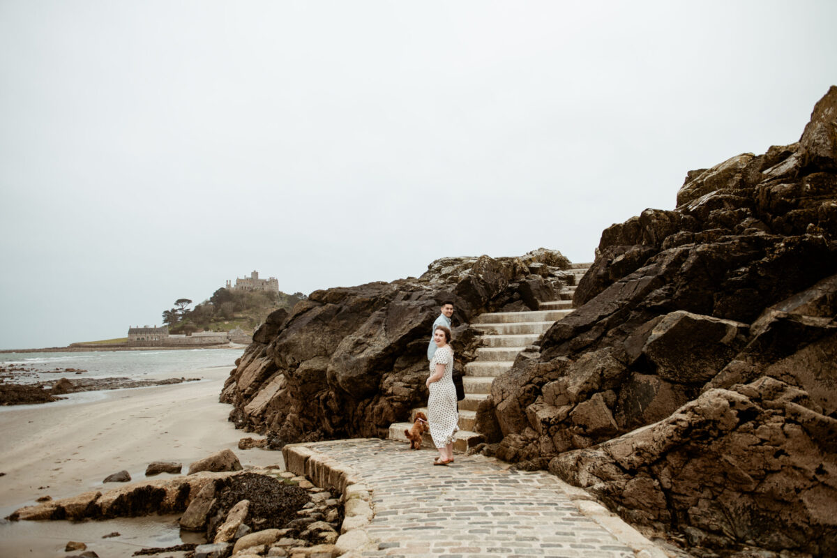 Beth and Tay walking up the stone steps at Marazion beach with St Michael's Mount in the background, their dog at their heels — Cornwall engagement photography