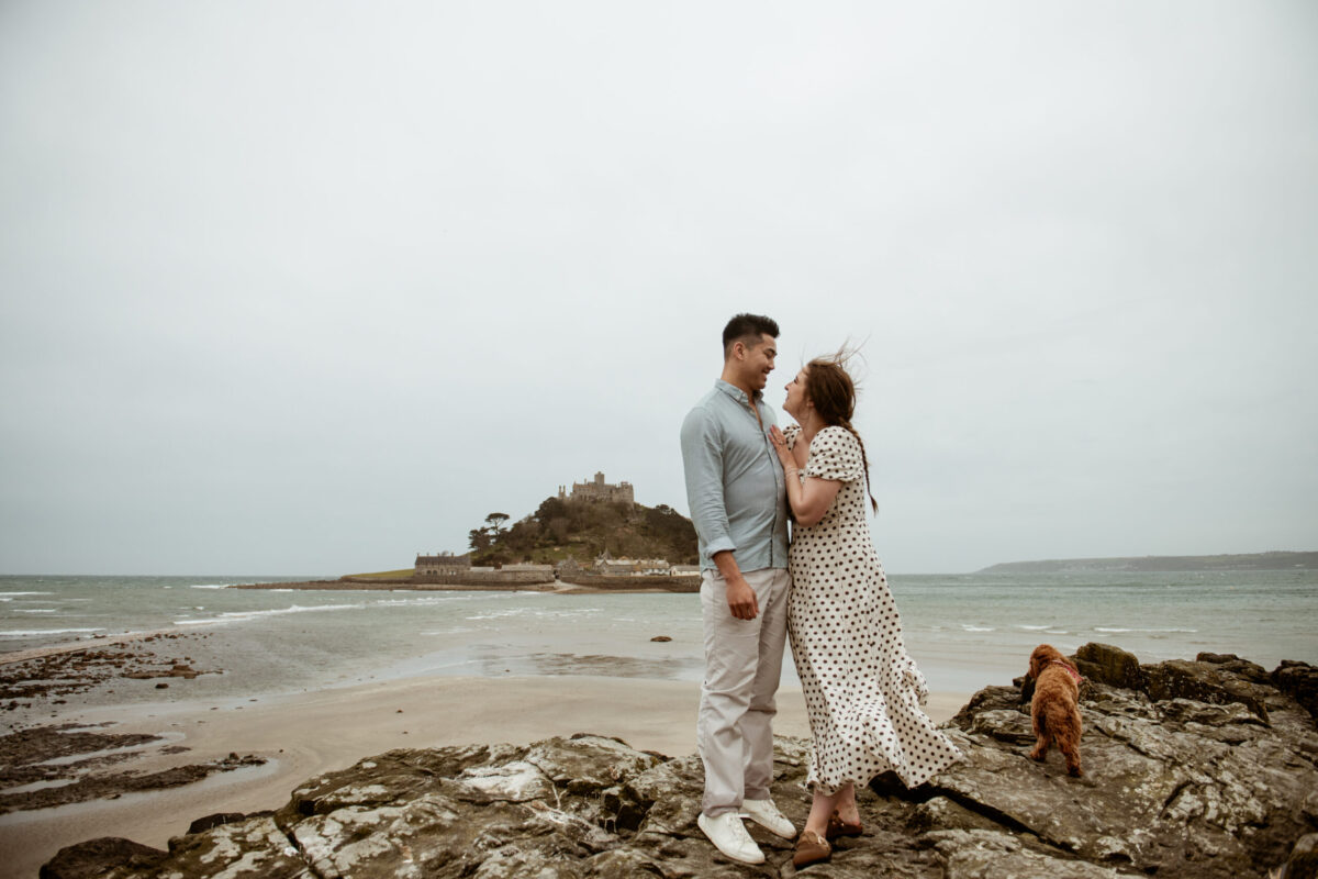 Tay and Beth laughing on the rocks at Marazion beach with St Michael's Mount behind them and their dog — Cornwall engagement photographer
