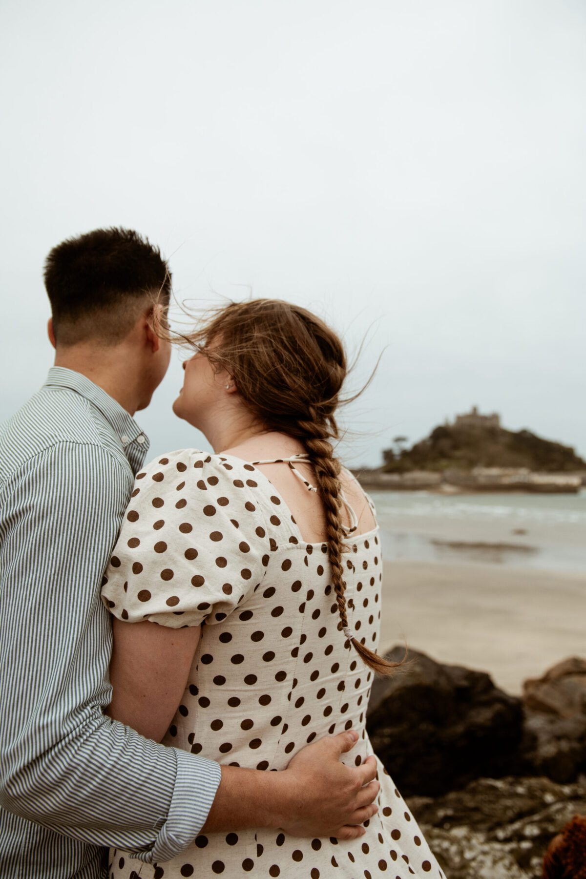 Tay holding Beth from behind as she looks toward St Michael's Mount, hair windswept, Marazion Cornwall