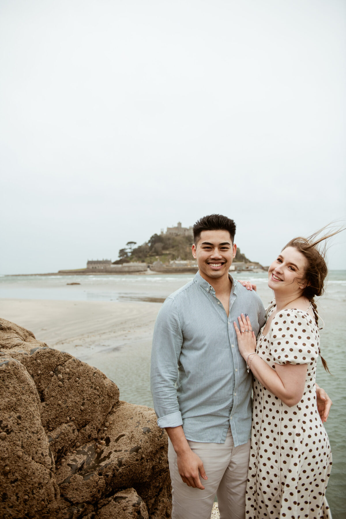 Tay and Beth smiling together on the rocks at Marazion, St Michael's Mount behind them, newly engaged — The Menagerie Lifestyle Photography