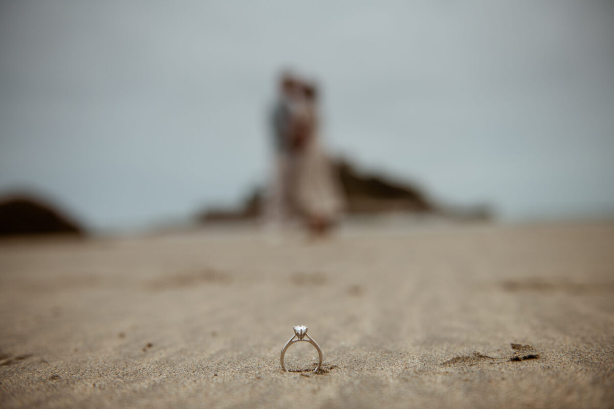 Diamond engagement ring standing in the sand at Marazion beach, couple softly blurred in the background